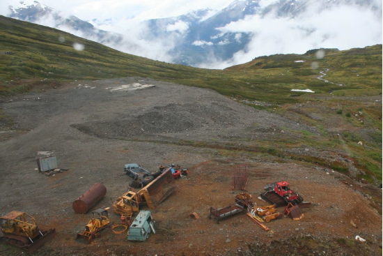 Discarded mining equipment lays on red soil in the foreground, with green hills and cloudy mountains in the background