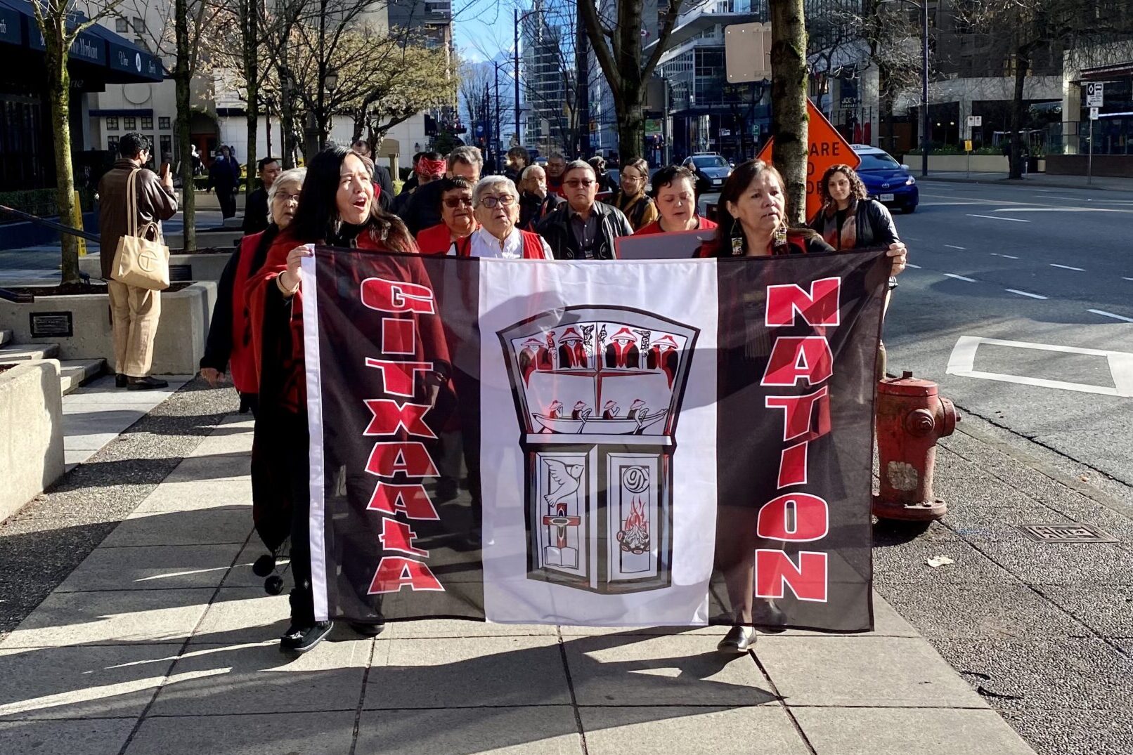 Three women lead a procession to the Vancouver Courthouse, holding the black, red and white flag of the Gitxaała Nation